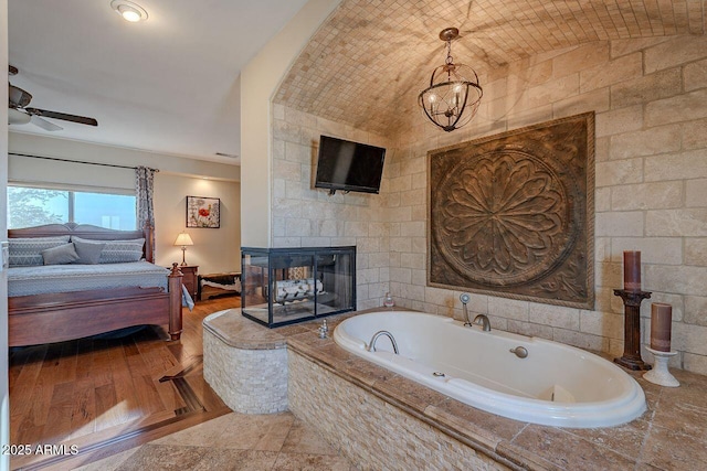 bathroom featuring a tiled fireplace, brick ceiling, ceiling fan with notable chandelier, and a relaxing tiled tub