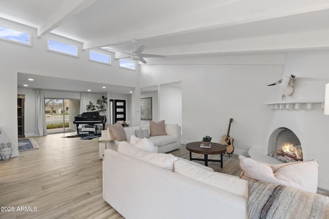 living room with beamed ceiling, light wood-style flooring, a warm lit fireplace, a high ceiling, and a ceiling fan