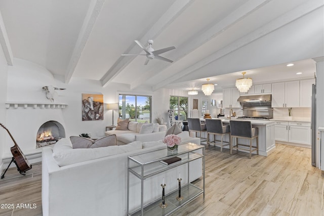 living area with light wood-style floors, a lit fireplace, vaulted ceiling with beams, and ceiling fan with notable chandelier