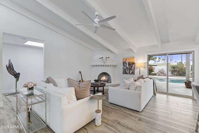 living area featuring ceiling fan, a lit fireplace, light wood-style flooring, and vaulted ceiling with beams