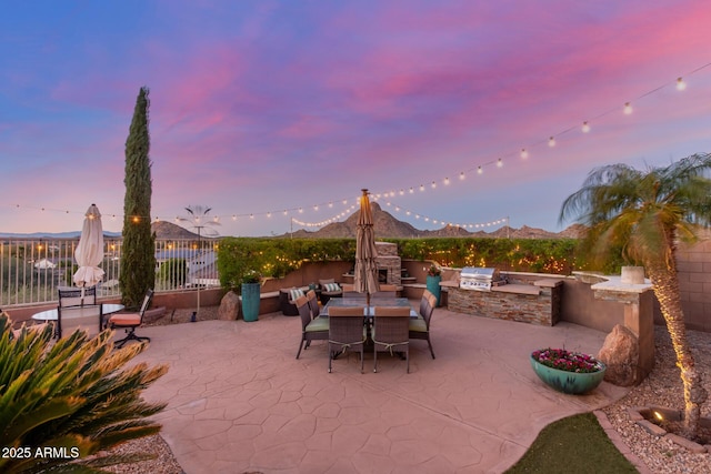 patio terrace at dusk with area for grilling, fence, a mountain view, exterior kitchen, and outdoor dining area