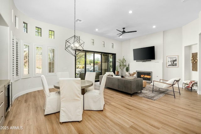 dining area featuring light wood finished floors, a high ceiling, recessed lighting, a glass covered fireplace, and ceiling fan with notable chandelier