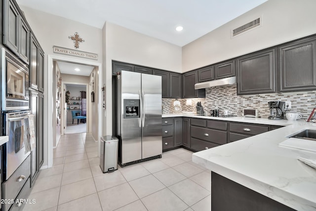 kitchen featuring light stone countertops, decorative backsplash, dark brown cabinets, light tile patterned flooring, and appliances with stainless steel finishes