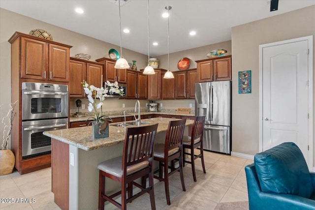 kitchen featuring an island with sink, light stone counters, hanging light fixtures, stainless steel appliances, and a sink