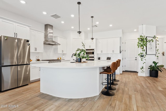 kitchen featuring appliances with stainless steel finishes, wall chimney exhaust hood, a kitchen island with sink, tasteful backsplash, and light countertops