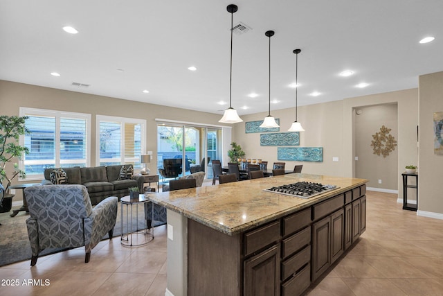 kitchen featuring stainless steel gas stovetop, hanging light fixtures, light tile patterned floors, dark brown cabinets, and light stone counters