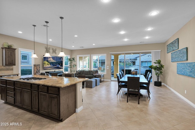 kitchen featuring pendant lighting, stainless steel gas stovetop, a center island, light stone countertops, and dark brown cabinets