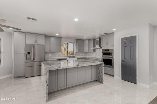 kitchen featuring light stone counters, wall chimney exhaust hood, a center island, gray cabinetry, and appliances with stainless steel finishes