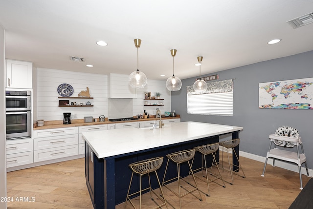 kitchen featuring stainless steel double oven, a kitchen island with sink, light hardwood / wood-style flooring, white cabinets, and hanging light fixtures