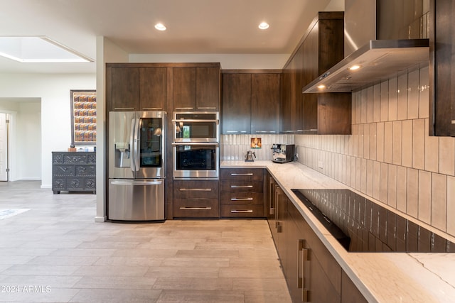 kitchen with wall chimney exhaust hood, dark brown cabinetry, light stone counters, appliances with stainless steel finishes, and decorative backsplash