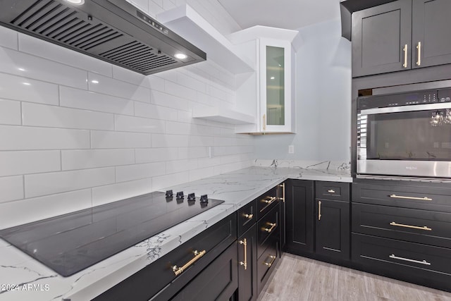 kitchen with wall chimney exhaust hood, light stone countertops, white cabinets, black electric cooktop, and stainless steel oven