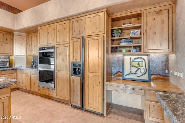 kitchen with built in desk, light brown cabinets, dark stone counters, light tile patterned floors, and stainless steel double oven