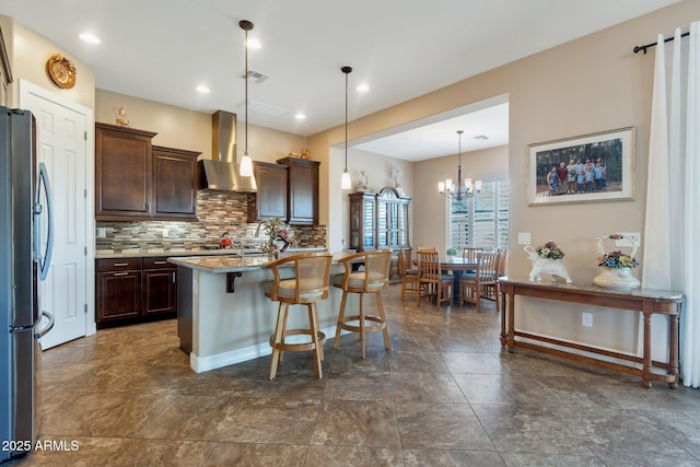 kitchen with backsplash, freestanding refrigerator, dark brown cabinetry, wall chimney range hood, and a kitchen bar