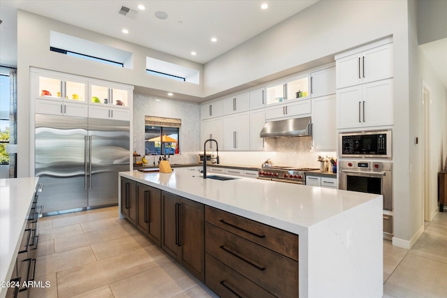 kitchen featuring sink, a towering ceiling, built in appliances, an island with sink, and white cabinets