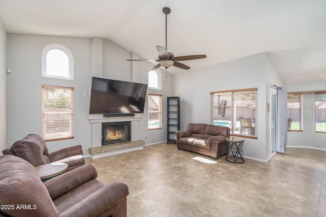 living room featuring a ceiling fan, baseboards, a warm lit fireplace, and high vaulted ceiling