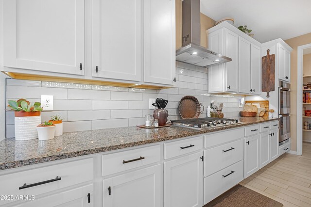 kitchen with stone counters, backsplash, white cabinetry, wall chimney exhaust hood, and appliances with stainless steel finishes