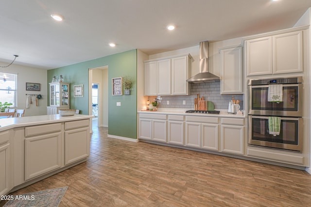 kitchen featuring wall chimney range hood, light countertops, decorative backsplash, light wood-style floors, and stainless steel appliances