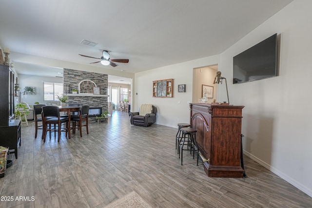 dining space featuring visible vents, ceiling fan, baseboards, a stone fireplace, and wood finished floors