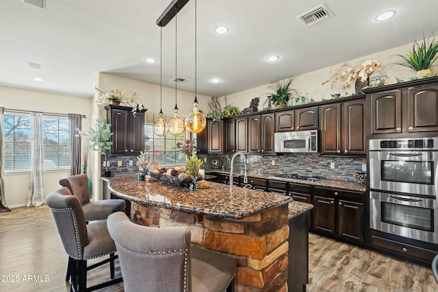 891 W Palo Brea Dr, Litchfield Park, AZ 85340 - kitchen featuring dark brown cabinets, dark stone counters, a kitchen island with sink, appliances with stainless steel finishes, and decorative light fixtures