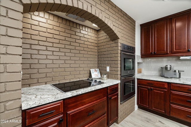 kitchen with black electric cooktop, light stone countertops, stainless steel double oven, brick wall, and light hardwood / wood-style flooring