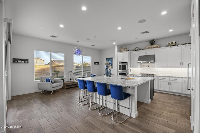 kitchen with a breakfast bar, double oven, an island with sink, white cabinets, and light stone counters