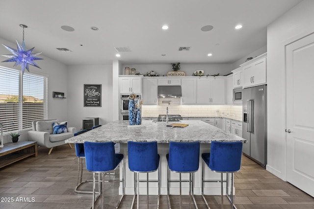 kitchen with stainless steel appliances, white cabinetry, a kitchen island with sink, and light stone counters