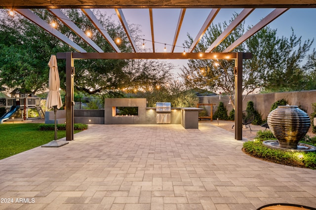 patio terrace at dusk featuring a playground, an outdoor kitchen, a pergola, and grilling area