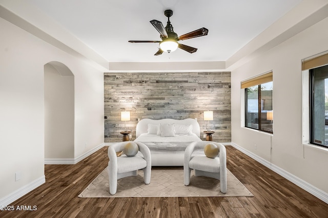 bedroom featuring ceiling fan, arched walkways, an accent wall, baseboards, and wood finished floors