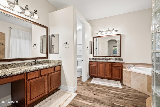 bathroom featuring a sink, toilet, two vanities, wood finished floors, and a garden tub