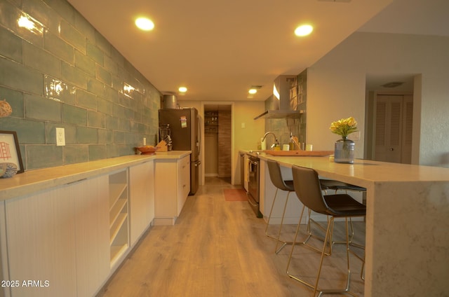 bar with stainless steel refrigerator, white cabinetry, wall chimney exhaust hood, and light wood-type flooring