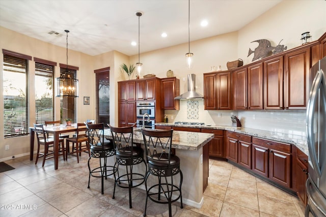 kitchen featuring appliances with stainless steel finishes, hanging light fixtures, a kitchen island with sink, light stone countertops, and wall chimney range hood