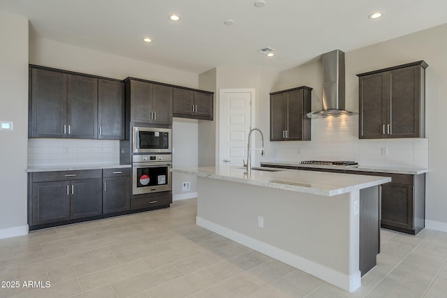 kitchen with stainless steel appliances, wall chimney range hood, a kitchen island with sink, dark brown cabinets, and sink