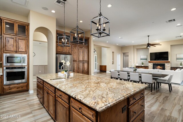 kitchen featuring ceiling fan with notable chandelier, stainless steel appliances, sink, a stone fireplace, and a center island with sink