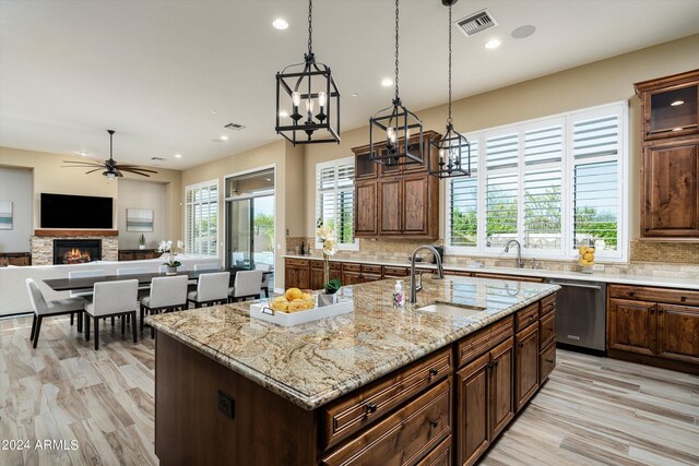 kitchen featuring ceiling fan with notable chandelier, a stone fireplace, backsplash, a kitchen island with sink, and stainless steel dishwasher