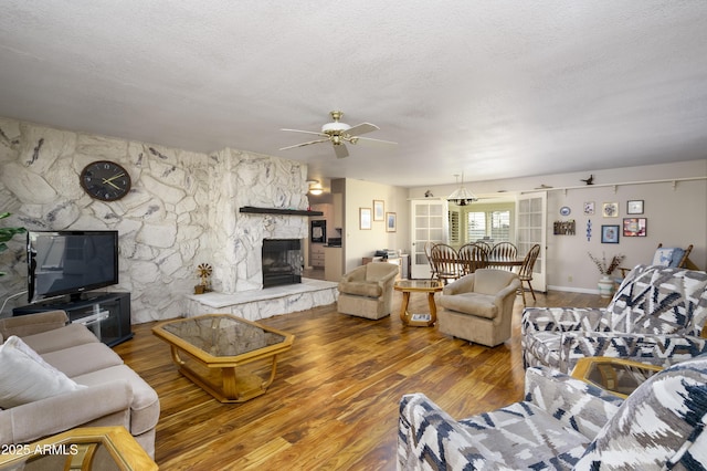 living room with a textured ceiling, a stone fireplace, wood finished floors, and a ceiling fan