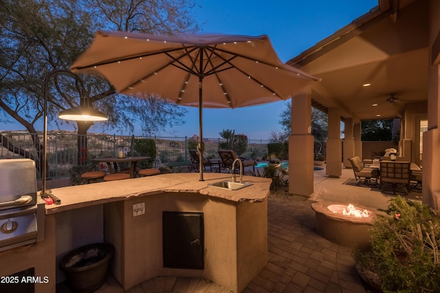 patio terrace at dusk with ceiling fan, sink, an outdoor kitchen, and a fire pit