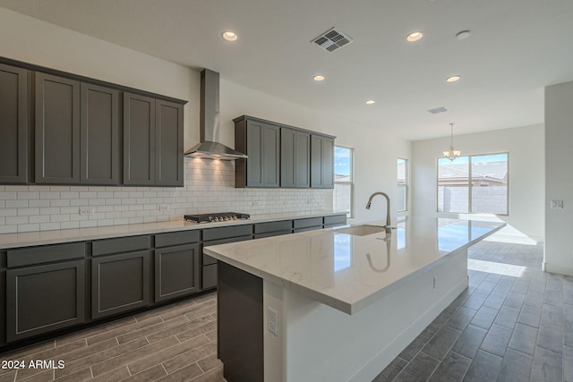 kitchen with wall chimney exhaust hood, sink, a chandelier, stainless steel gas stovetop, and an island with sink