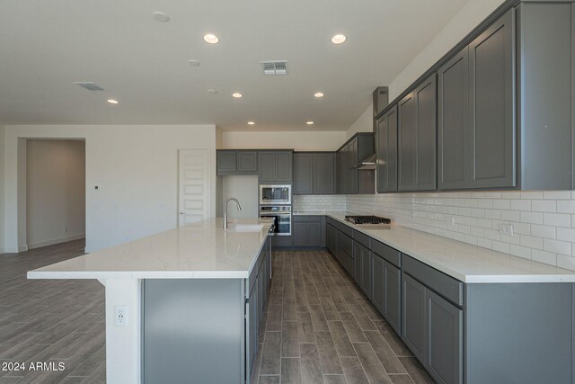 kitchen featuring decorative backsplash, gray cabinetry, stainless steel appliances, dark hardwood / wood-style floors, and an island with sink