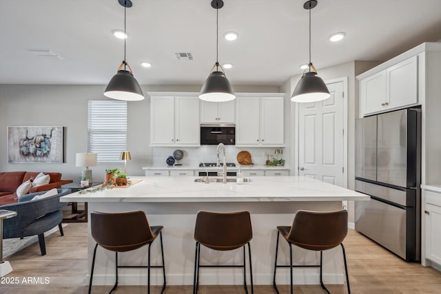 kitchen with white cabinets, hanging light fixtures, stainless steel refrigerator, and an island with sink