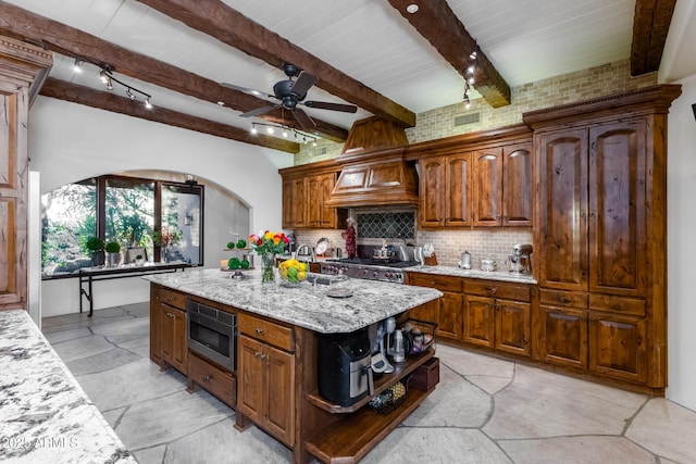 kitchen with beam ceiling, light stone countertops, stainless steel microwave, backsplash, and a center island with sink