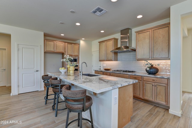 kitchen with wall chimney range hood, a kitchen island with sink, light hardwood / wood-style flooring, sink, and light stone countertops