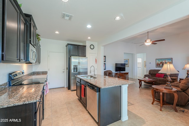 kitchen featuring stainless steel appliances, an island with sink, ceiling fan, sink, and light stone counters