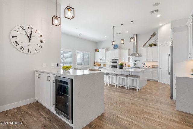 kitchen with white cabinets, wall chimney range hood, hanging light fixtures, light wood-type flooring, and beverage cooler