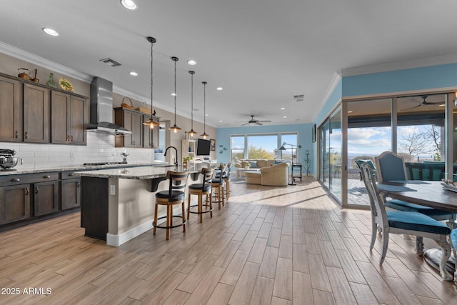 kitchen featuring pendant lighting, an island with sink, a breakfast bar area, light stone counters, and wall chimney range hood