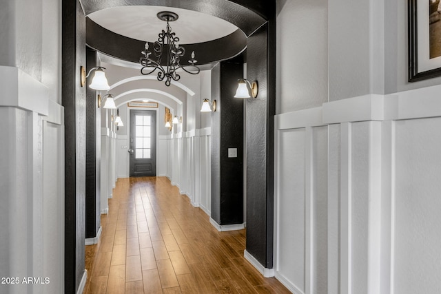 hall with arched walkways, a wainscoted wall, wood finished floors, a decorative wall, and a notable chandelier