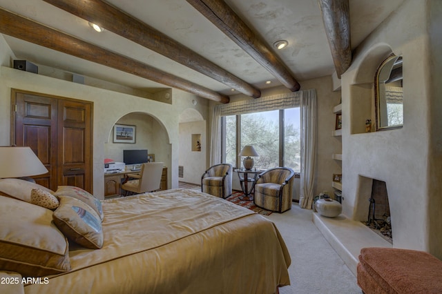 bedroom with beam ceiling, a fireplace with raised hearth, arched walkways, and carpet floors