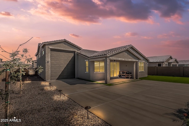 back of property at dusk featuring stucco siding, a tile roof, fence, concrete driveway, and a garage