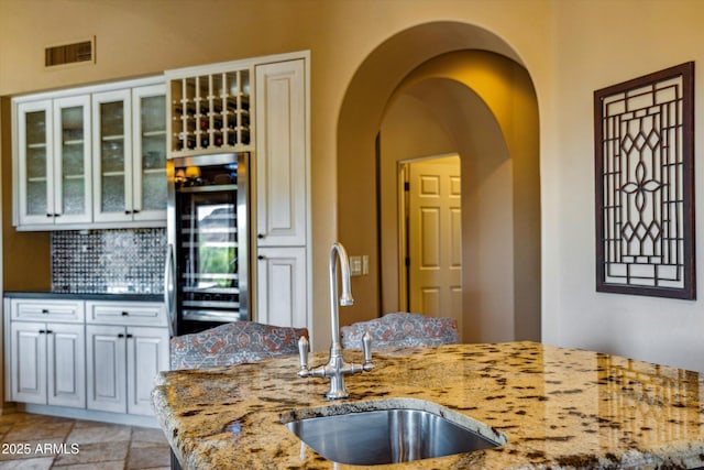 kitchen with a sink, beverage cooler, light stone countertops, arched walkways, and glass insert cabinets