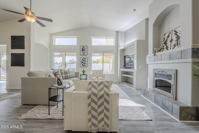 living area with high vaulted ceiling, a tiled fireplace, heating unit, wood finished floors, and baseboards