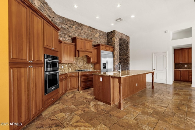 kitchen featuring sink, backsplash, stainless steel appliances, light stone countertops, and a center island with sink